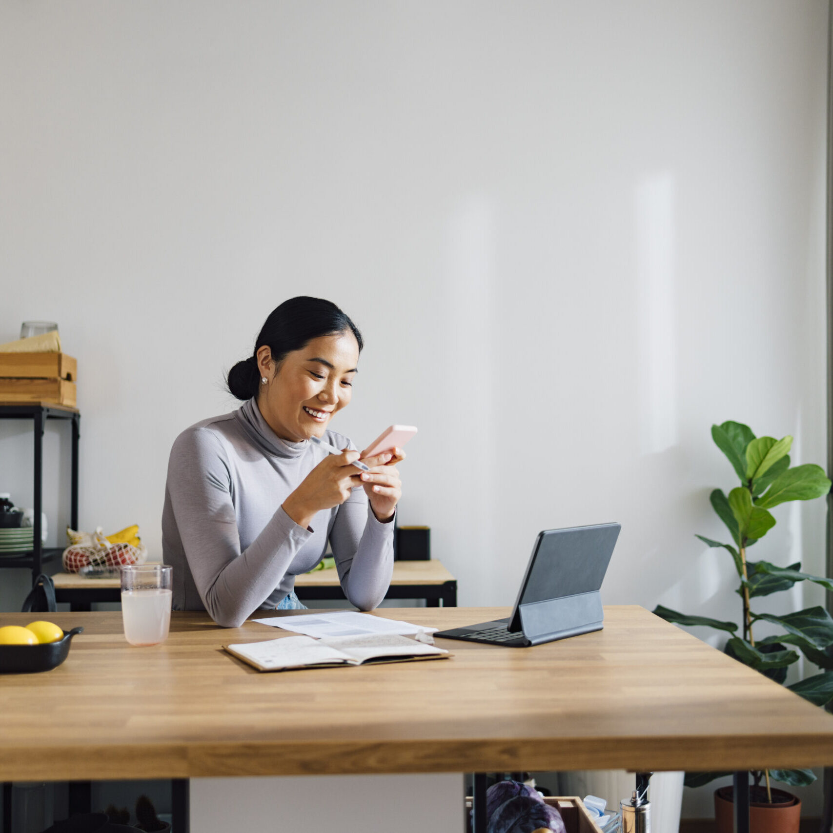 Beautiful Asian businesswoman using digital tablet and notebook while typing text message on her mobile phone at kitchen desk.