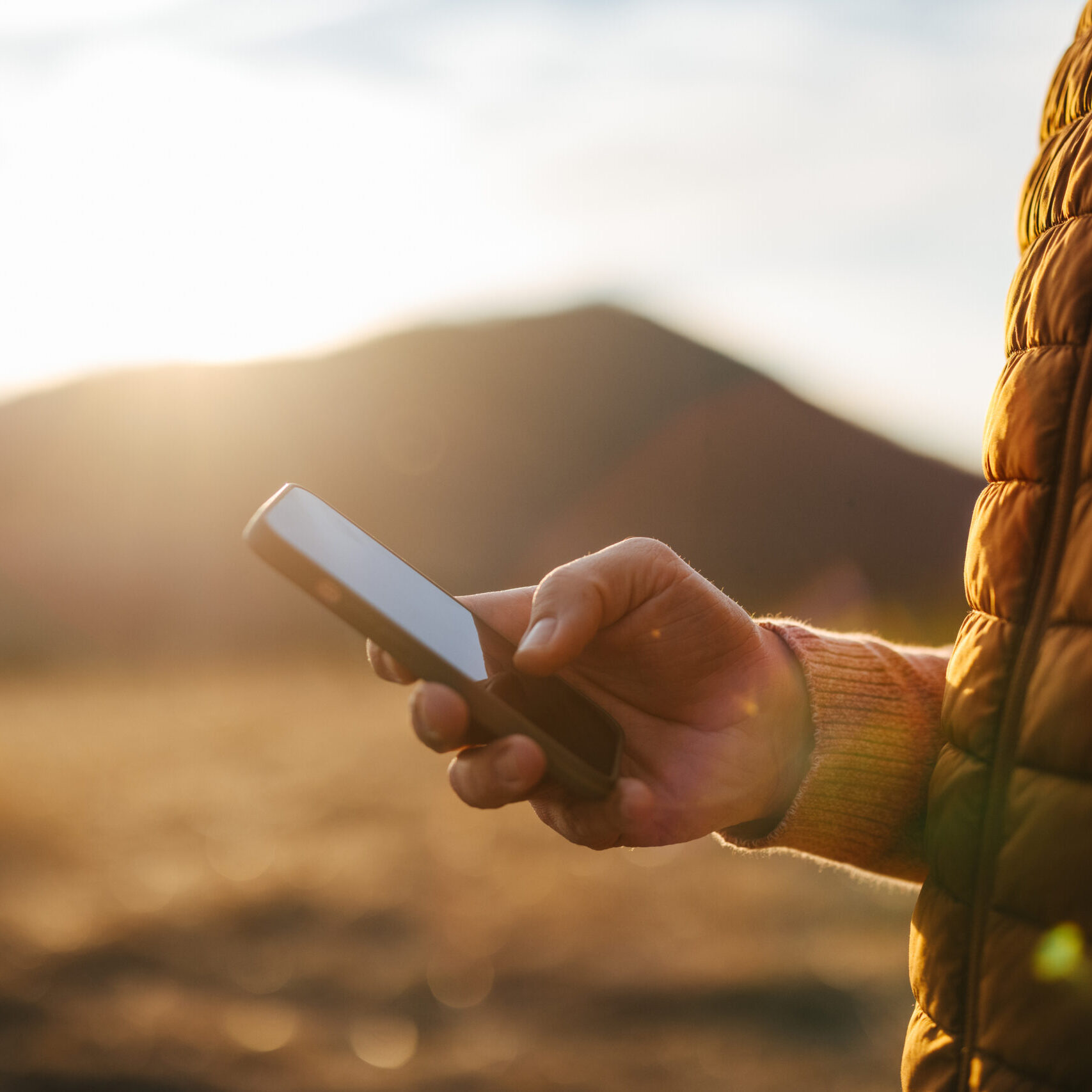 Close-up of a person holding a smartphone in the mountains during sunset, enjoying a relaxing vacation. The warm light adds a cozy feeling to the outdoor scene.