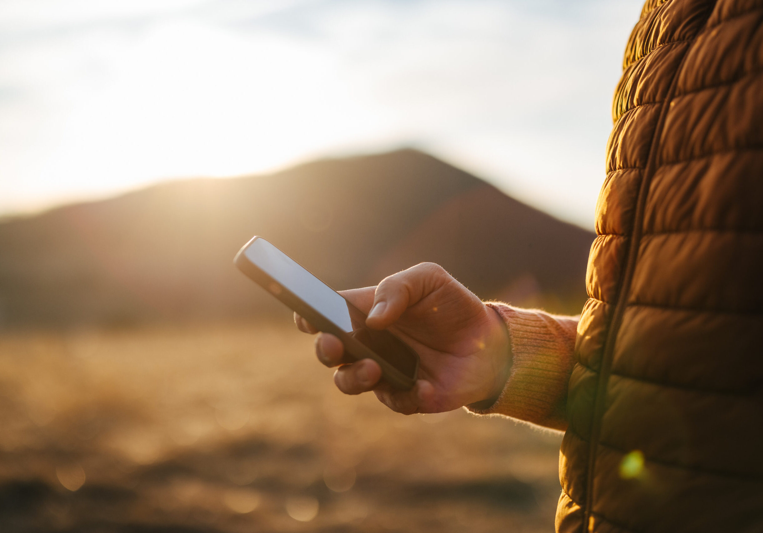 Close-up of a person holding a smartphone in the mountains during sunset, enjoying a relaxing vacation. The warm light adds a cozy feeling to the outdoor scene.
